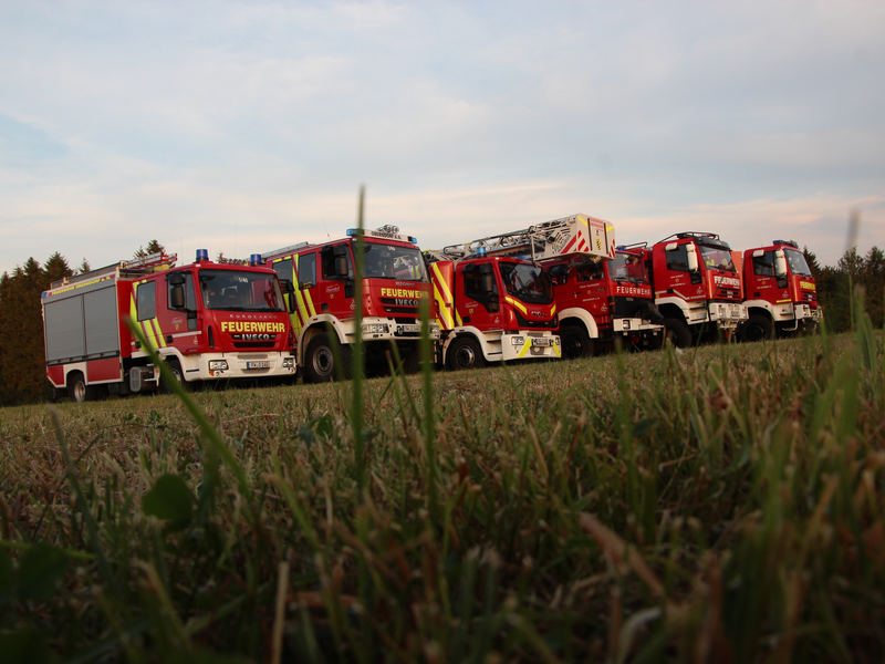Sechs verschiedene Feuerwehrautos stehen nebeneinander aufgereiht auf einer Wiese. Perspektive aus der Seiten- und Vorderansicht. Alle sechs sind verschiedene Feuerwehrfahrzeuge. 