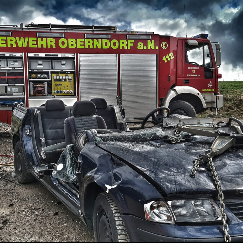 Ein völlig schwarz abgebranntes Auto ohne Dach im Vordergrund. Im Hintergrund steht ein Feuerwehrauto. Der Himmel ist voller dunkler Wolken. 