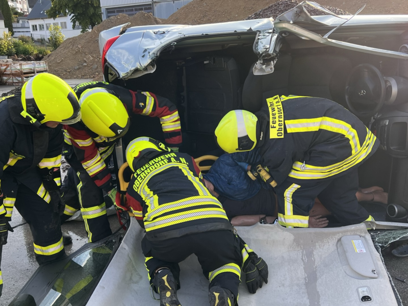 Vier Feuerwehr Personen retten eine Übungspuppe aus einem auf der Seite liegenden Auto welches kein Dach mehr hat. 