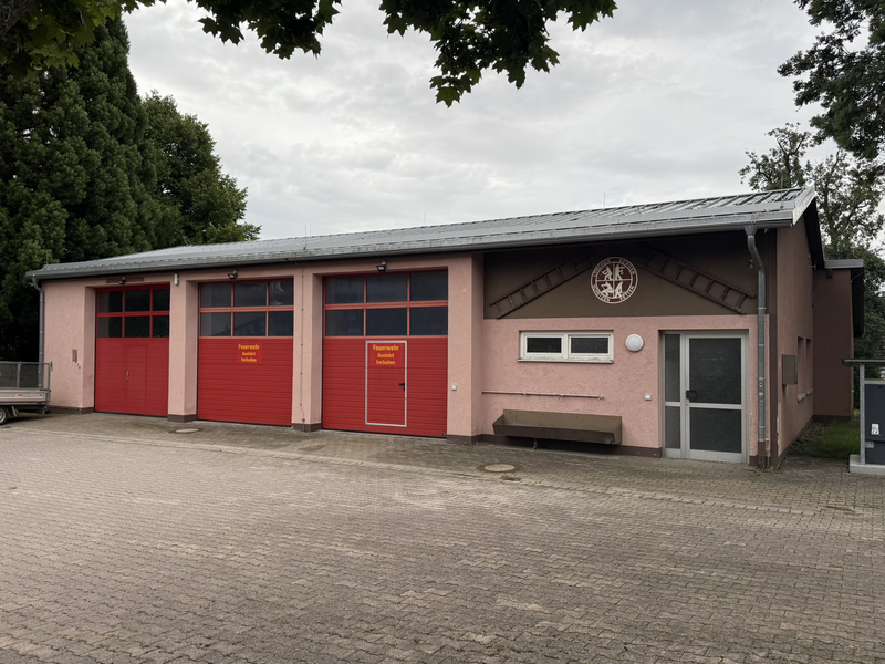 Feuerwehrhaus Hochmössingen mit drei roten Garagen für die Fahrzeuge. Fassade ist rosa gestrichen. Neben der Türe ist eine Wanne an der Wand angebracht. Über der Türe sind zwei Leitern und ein Logo zu sehen. 