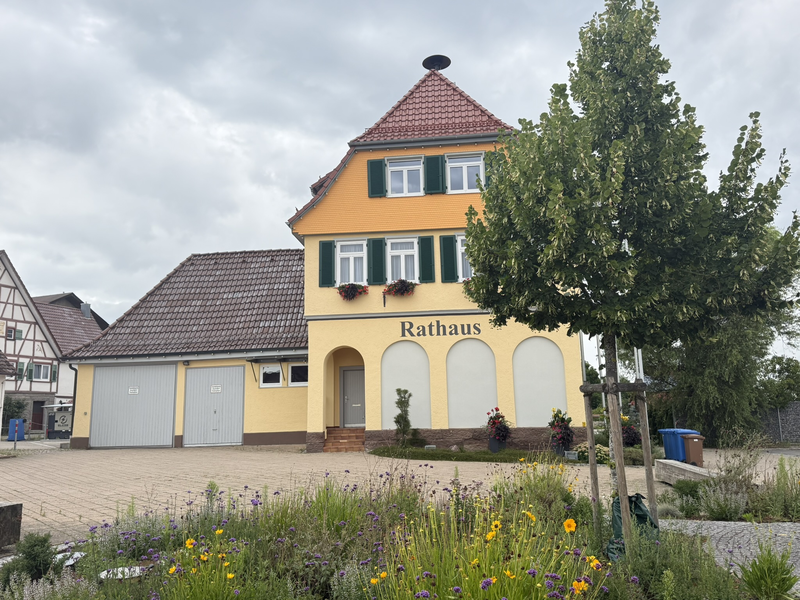 Feuerwehrhaus Boll mit einem großen und einem kleineren grauen Gagragentor. Haus hat eine gelbe Fassade und die Fenster grüne Fensterläden. Ein Baum ist im Vordergrund zu sehen. 