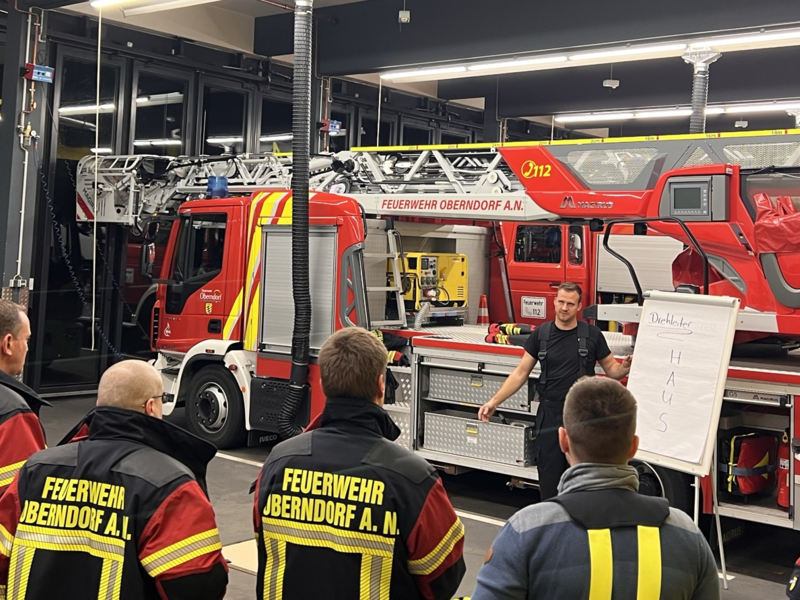 Taktische Drehleiterausbildung im Rahmen des Übungsbetriebs. 11 männliche Personen im Halbkreis mit Rückenansicht. Der Schulungsleiter steht vor einem Feuerwehrauto, neben ihm steht ein Flip-Chart. 