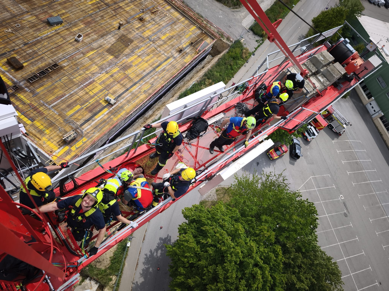 11 Menschen auf einem Kran bei einem Rettungseinsatz. Sicht von ob. Alle in Schutzkleidung. Kran steht über einem Parkplatz