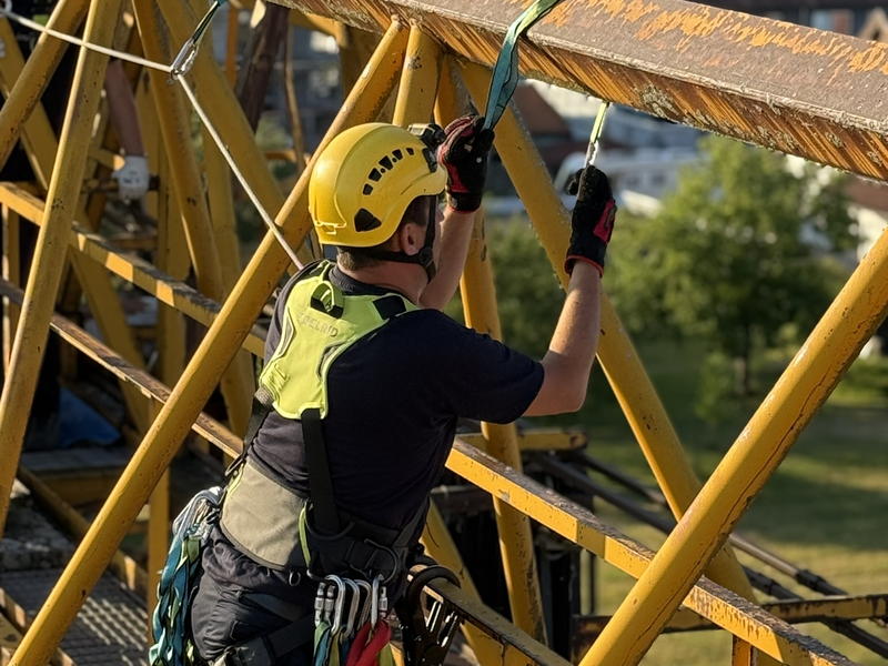 Mann mit Schutzkleidung und gelben Helm auf der steht auf der Außenseiten eines Krans und ist dort abgesichert. 