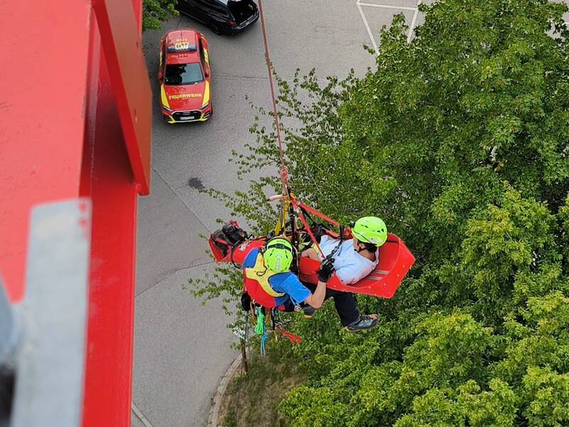 Zwei Männer werden von einem Kran abgeseilt. Einer liegt in einer Transportliege der andere hängt abgesichert am Seil. Sicht von oben. 
