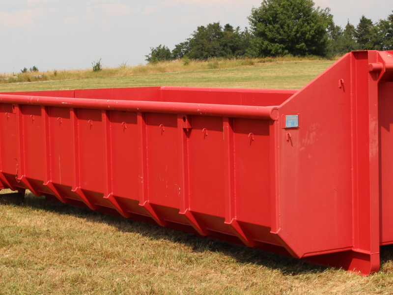 Roter Muldencontainer steht auf einer Wiese. Im Hintergrund ist ein Wald zu sehen. 