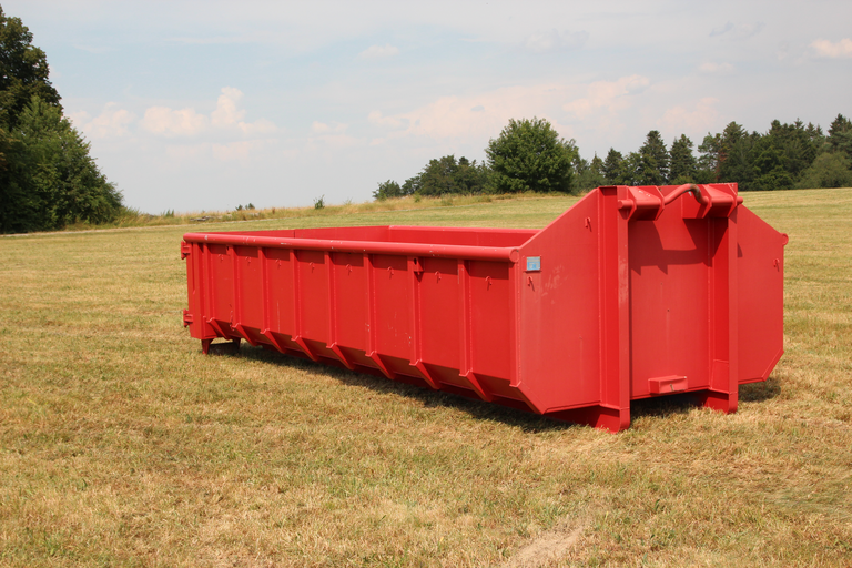 Roter Muldencontainer steht auf einer Wiese. Im Hintergrund ist ein Wald zu sehen. 