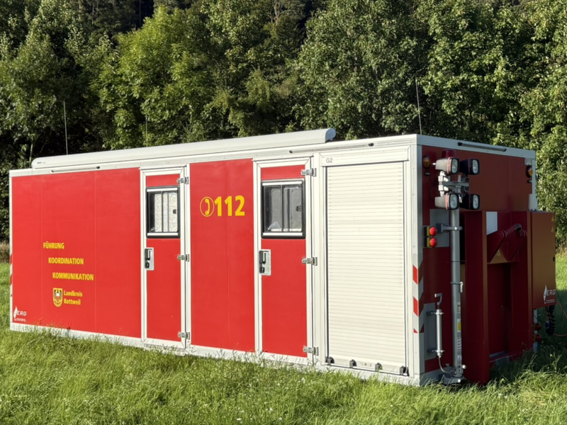 Roter Container mit zwei Türen und Fenster steht auf einer Wiese. Gelber Schriftzug Führung, Koordination, Kommunikation. Notrufnummer 112 ist ebenfalls abgebildet. 
