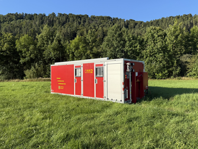 Roter Container mit zwei Türen und Fenster steht auf einer Wiese. Gelber Schriftzug Führung, Koordination, Kommunikation. Notrufnummer 112 ist ebenfalls abgebildet. 