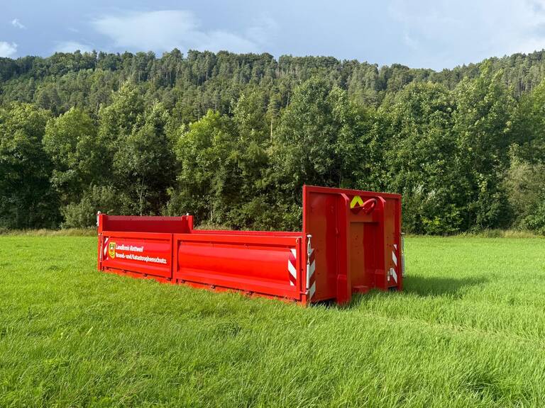 Eine rote Pritsche steht auf einer Wiese. Im Hintergrund ist Wald zu sehen. 