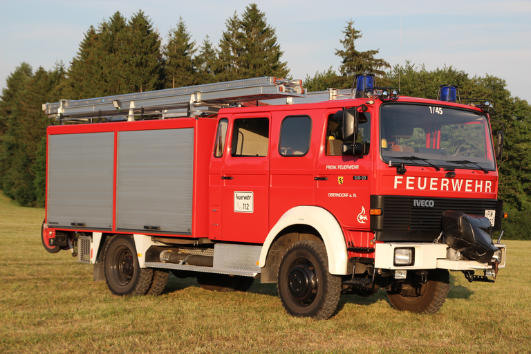 Feuerwehr-Löschfahrzeug steht auf einer Wiese mit Wald im Hintergrund. Löschfahrzeug mit zwei Rolltoren an der Seite und Leiter auf dem Dach. Auf der Frontscheibe steht Fahrzeugbezeichnung 1/45