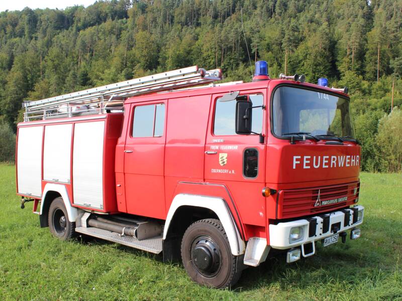 Älteres rotes Feuerwehrauto in Seitenansicht stehend auf einer Wiese mit Wald im Hintergrund. Das Auto hat drei graue Rolltore an der Seite und das Logo der Stadt Oberndorf auf der Beifahrertür. 