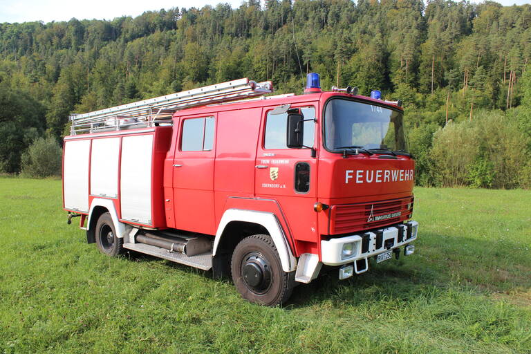 Älteres rotes Feuerwehrauto in Seitenansicht stehend auf einer Wiese mit Wald im Hintergrund. Das Auto hat drei graue Rolltore an der Seite und das Logo der Stadt Oberndorf auf der Beifahrertür. 