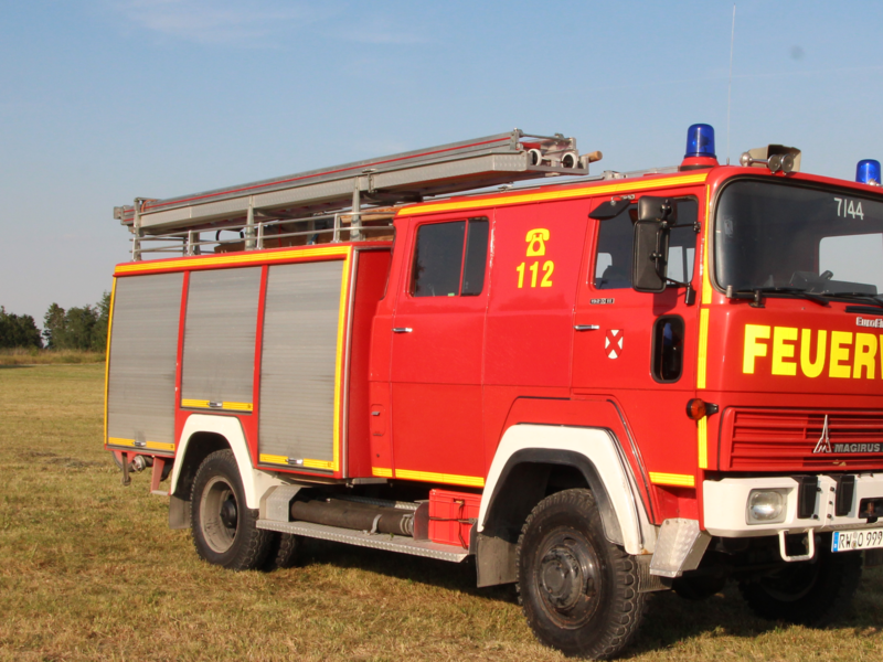 Älteres rot/gelbes Feuerwehrauto mit drei grauen Rolltoren an der Seite. Auf dem Dach ist eine Leiter angebracht. 
