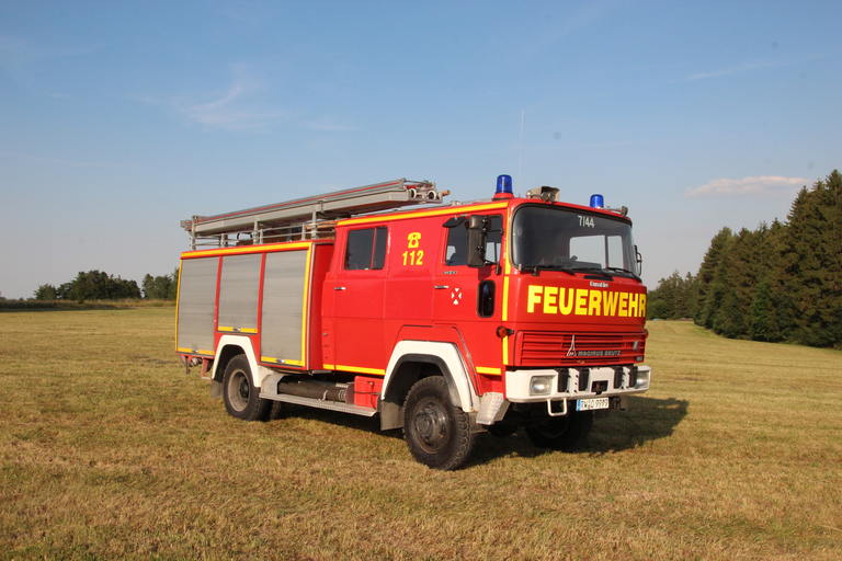 Älteres rot/gelbes Feuerwehrauto mit drei grauen Rolltoren an der Seite. Auf dem Dach ist eine Leiter angebracht. 
