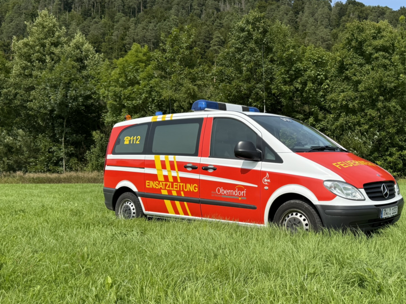 Feuerwehr Kleinbus steht seitlich auf einer Wiese. Marke Mercedes Benz. Farbe Feuerwehrrot und Neongelb. 