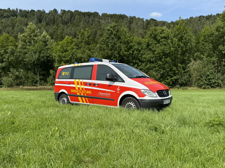 Feuerwehr Kleinbus steht seitlich auf einer Wiese. Marke Mercedes Benz. Farbe Feuerwehrrot und Neongelb. 