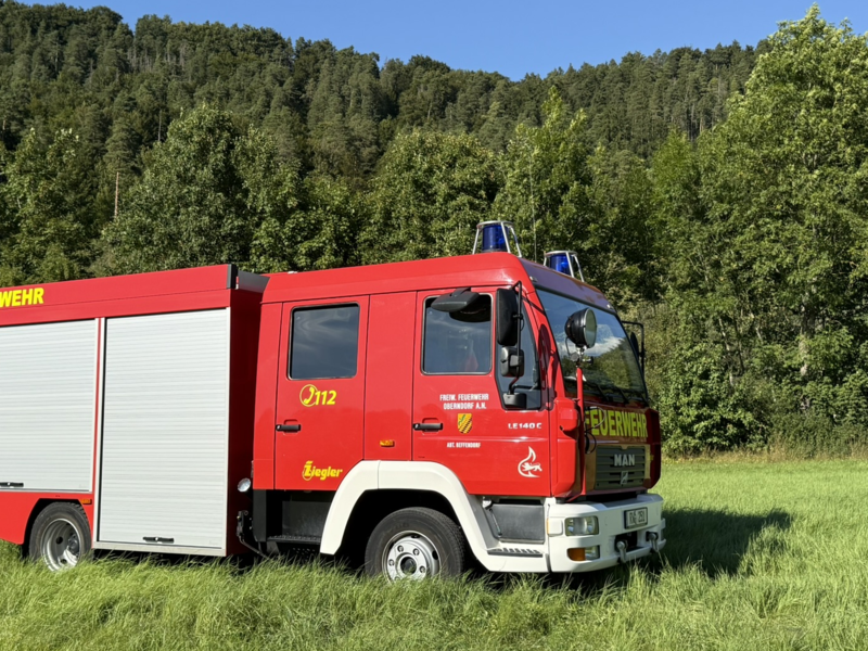 Rotes Feuerwehrauto mit zwei grauen Rolltoren an der Seite. Schriftzüge der Feuerwehr und der Stadt Oberndorf sind gelb abgebildet. Auf dem Dach der Fahrerkabine sind zwei blaue Warnlichter angebracht. 