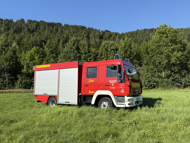 Rotes Feuerwehrauto mit zwei grauen Rolltoren an der Seite. Schriftzüge der Feuerwehr und der Stadt Oberndorf sind gelb abgebildet. Auf dem Dach der Fahrerkabine sind zwei blaue Warnlichter angebracht. 