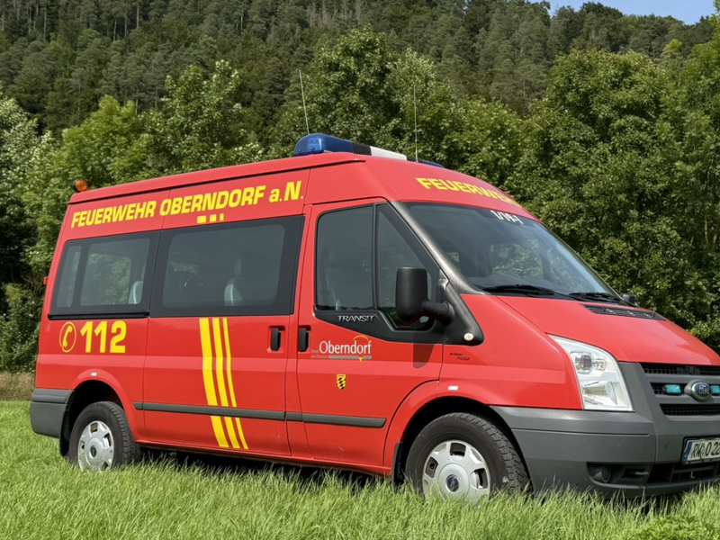 Rot/Gelber Kleintransporter mit mehreren Sitzplätzen. Auf der Seite gibt es einen Schriftzug mit Feuerwehr Oberndorf a.N. und der Telefonnummer 112. Das Fahrzeug steht auf einer Wiese. Im Hintergrund ist ein Wald zu sehen. 