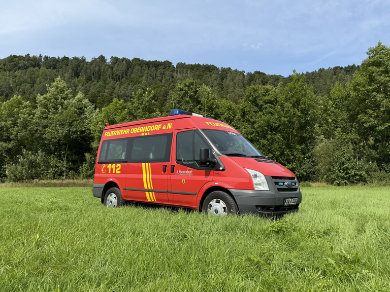 Rot/Gelber Kleintransporter mit mehreren Sitzplätzen. Auf der Seite gibt es einen Schriftzug mit Feuerwehr Oberndorf a.N. und der Telefonnummer 112. Das Fahrzeug steht auf einer Wiese. Im Hintergrund ist ein Wald zu sehen. 