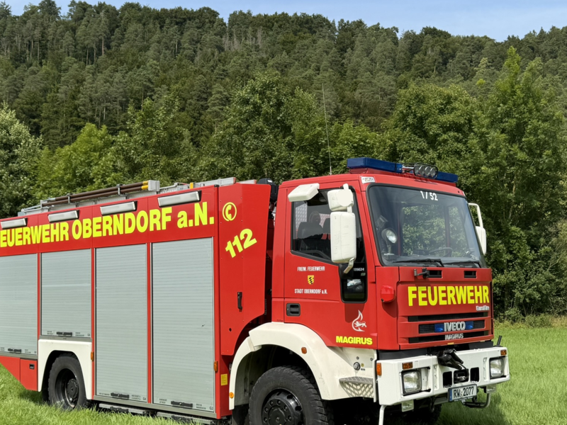 Feuerwehrauto mit Seitansicht steht auf einer Wiese mit Wald im Hintergrund. Das Feuerwehrauto hat vier Rolltore. Gelber Schriftzug mit "Feuerwehr Oberndorf a. N." und der Notrufnummer "112" ist seitliche abgedruckt. 