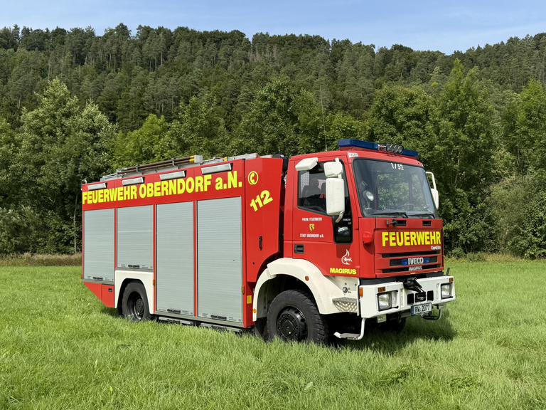 Feuerwehrauto mit Seitansicht steht auf einer Wiese mit Wald im Hintergrund. Das Feuerwehrauto hat vier Rolltore. Gelber Schriftzug mit "Feuerwehr Oberndorf a. N." und der Notrufnummer "112" ist seitliche abgedruckt. 