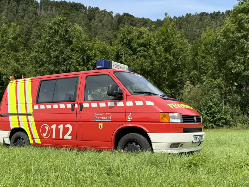 Feuerwehr Kleinbus steht seitlich auf einer Wiese mit Wald im Hintergrund. Hinter der Beifahrertür ist eine Schiebetür angebracht. Der Bus ist rot gelb lackiert. Das Logo der Stadt Oberndorf sowie die Notrufnummer 112 sind seitlich aufgebracht. 