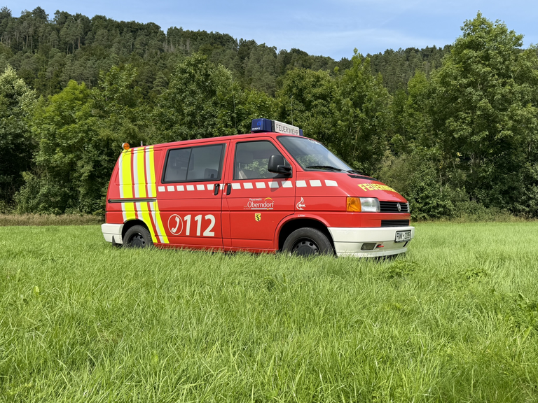 Feuerwehr Kleinbus steht seitlich auf einer Wiese mit Wald im Hintergrund. Hinter der Beifahrertür ist eine Schiebetür angebracht. Der Bus ist rot gelb lackiert. Das Logo der Stadt Oberndorf sowie die Notrufnummer 112 sind seitlich aufgebracht. 