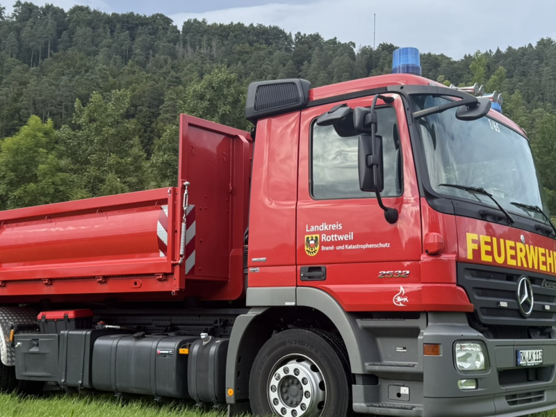 Wechselladerfahrzeug mit aufgesattelter Pritsche steht auf grüner Wiese vor einem grünen Wald.