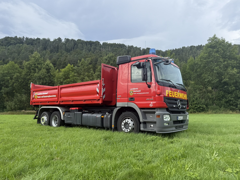 Wechselladerfahrzeug mit aufgesattelter Pritsche steht auf grüner Wiese vor einem grünen Wald.