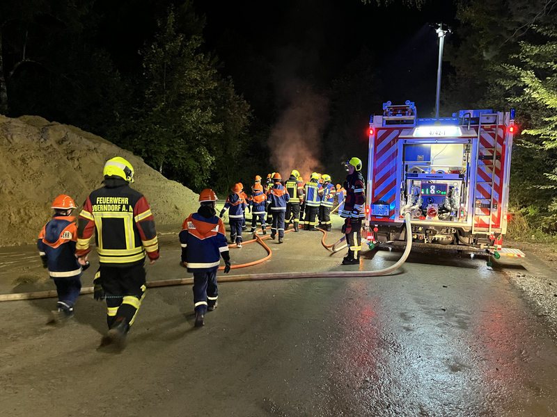 Im Dunkeln steht ein Feuerwehrauto mit ausgelassen Schläuchen am Rand. Viele junge Feuerwehr-Jugendliche und -Kinder nehmen an einer Übung teil. 
