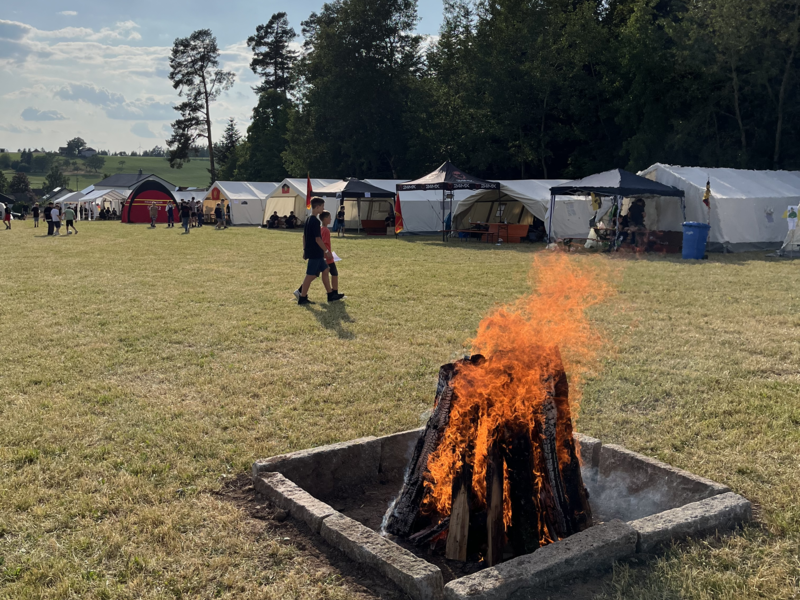 Offenes Lagerfeuer in der Mitte einer Wiese. Im Hintergrund sind viele Zelte vor dem Waldrand zu sehen.