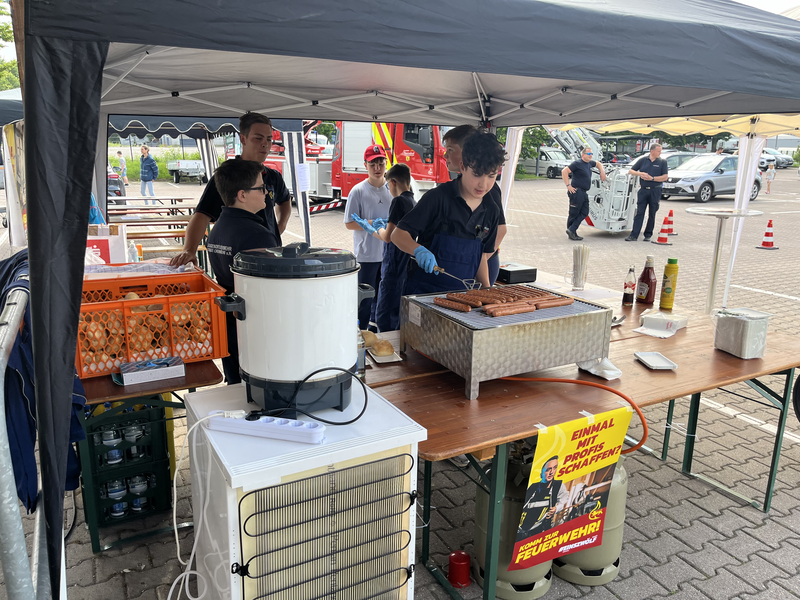Jugendliche stehen unter einem Zelt und grillen Würstchen und bieten Getränke zum verkauf auf. 