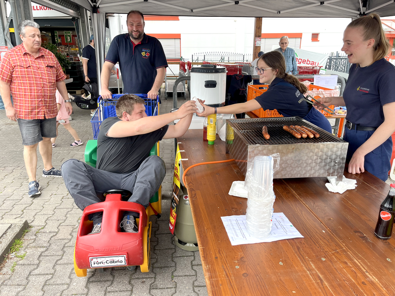 Zwei Mädchen in Feuerwehrbegleidung verkaufen gegrillte Würstchen. Ein Mann im Kinder-Einkaufswagen nimmt sein Brötchen lächelnd entgegen. 