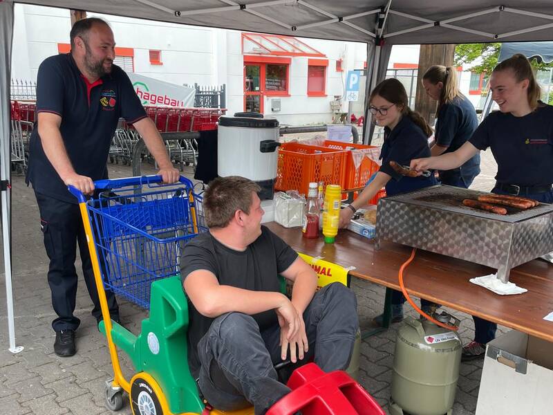 Ein Mann schiebt einen Kinder-Einkaufswagen in dem ein weiterer Mann sitzt. Dieser bestellt bei zwei Mädchen in Feuerwehruniform was zu essen. 