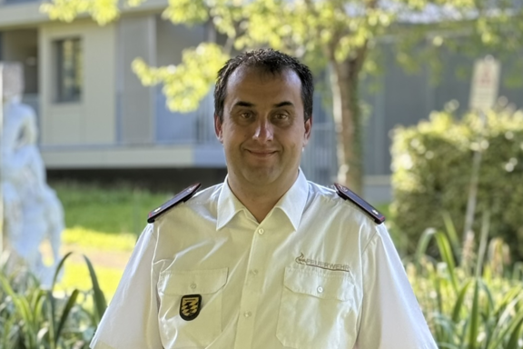 Eine männliche Person mit schwarzen Haaren steht auf einer Brücke und lächelt in die Kamera. Er hat ein weißes Feuerwehrhemd und schwarze Hose an. 