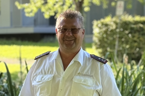 Eine männliche Person mit hellbraunen Haaren steht auf einer Brücke und lächelt in die Kamera. Er hat ein weißes Feuerwehrhemd und schwarze Hose an. 