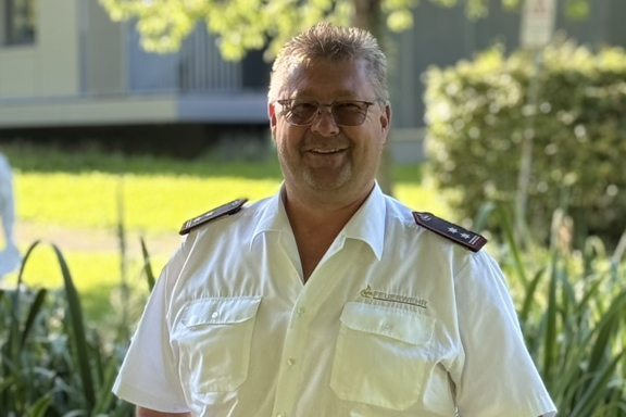 Eine männliche Person mit hellbraunen Haaren steht auf einer Brücke und lächelt in die Kamera. Er hat ein weißes Feuerwehrhemd und schwarze Hose an. 