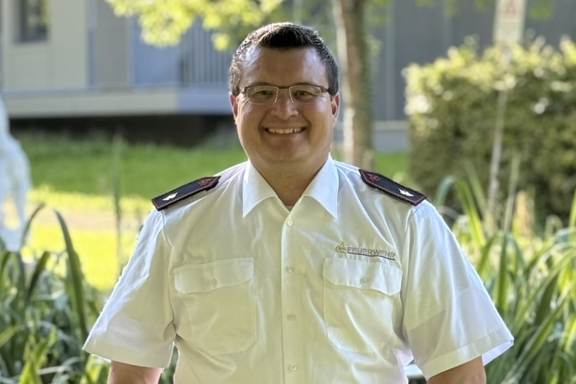 Eine männliche Person mit Brille steht auf einer Brücke und lächelt in die Kamera. Er hat ein weißes Feuerwehrhemd und schwarze Hose an. 