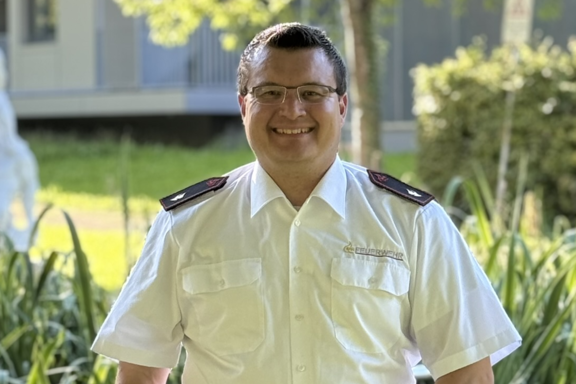 Eine männliche Person mit Brille steht auf einer Brücke und lächelt in die Kamera. Er hat ein weißes Feuerwehrhemd und schwarze Hose an. 