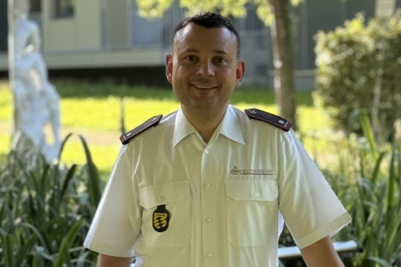 Eine männliche Person mit schwarzen Haaren steht auf einer Brücke und lächelt in die Kamera. Er hat ein weißes Feuerwehrhemd und schwarze Hose an. 