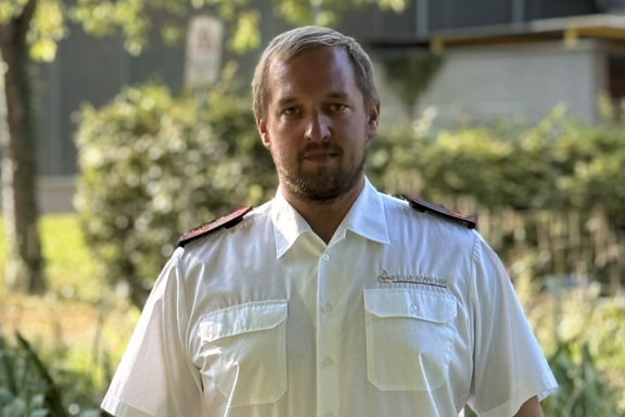 Eine männliche Person steht auf einer Brücke und lächelt in die Kamera. Er hat ein weißes Feuerwehrhemd und schwarze Hose an. 