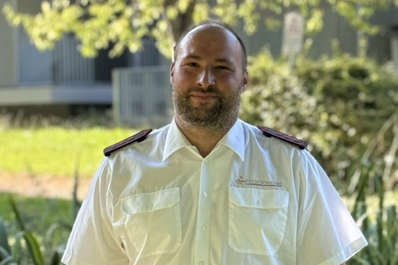 Eine männliche Person steht auf einer Brücke und lächelt in die Kamera. Er hat ein weißes Feuerwehrhemd und schwarze Hose an. 