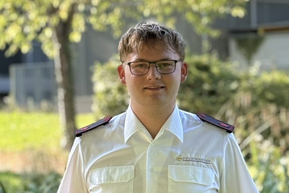 Eine männliche Person steht auf einer Brücke und lächelt in die Kamera. Er hat ein weißes Feuerwehrhemd und schwarze Hose an. 