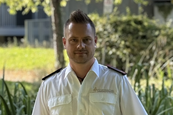 Eine männliche Person steht auf einer Brücke und lächelt in die Kamera. Er hat ein weißes Feuerwehrhemd und schwarze Hose an. 