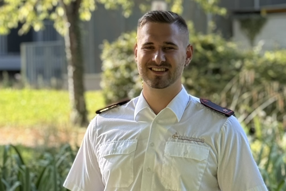 Eine männliche Person steht auf einer Brücke und lächelt in die Kamera. Er hat ein weißes Feuerwehrhemd und schwarze Hose an. 