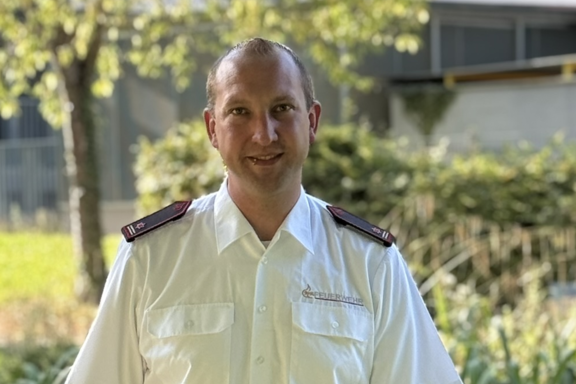 Eine männliche Person steht auf einer Brücke und lächelt in die Kamera. Er hat ein weißes Feuerwehrhemd und schwarze Hose an. 