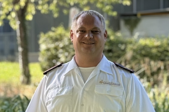 Eine männliche Person steht auf einer Brücke und lächelt in die Kamera. Er hat ein weißes Feuerwehrhemd und schwarze Hose an. 