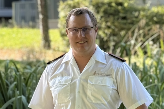 Eine männliche Person steht auf einer Brücke und lächelt in die Kamera. Er hat ein weißes Feuerwehrhemd und schwarze Hose an. 
